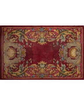 509-Alfombra en lana estilo Real Fábrica, c. 1940. Elegante diseño Carlos III a base de motivos dorados y guirnaldas de rosas sobre campo burdeos y rosa. Firmada J.G. Medidas: 285x315 cm.