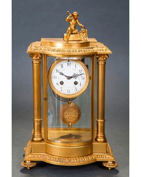 754-Reloj de sobremesa de estilo Imperio en bronce dorado y cristal, Francia, s. XIX. Rematado con pequeña escultura de bulto redondo, esfera blanca con decoración floral y numeración arábiga. Meca