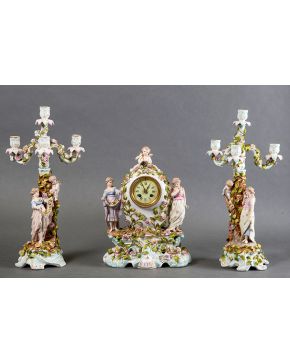 714-Reloj con guarnición de candelabros de cuatro luces en porcelana de Sajonía S.XIX. Fustes decorados con figuras de bulto redondo y flores aplicadas. Esfera blanca con numeración romana. Mecanismo 