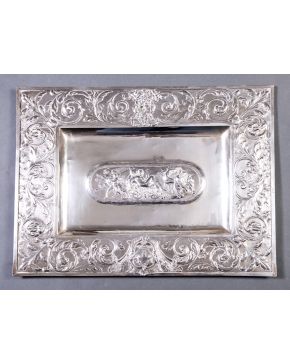 1006-Bandeja rectangular en plata de ley 916. Alero y centro cincelado con roleos vegetales y escena clásica.  Peso: 1.145 gr. Medidas